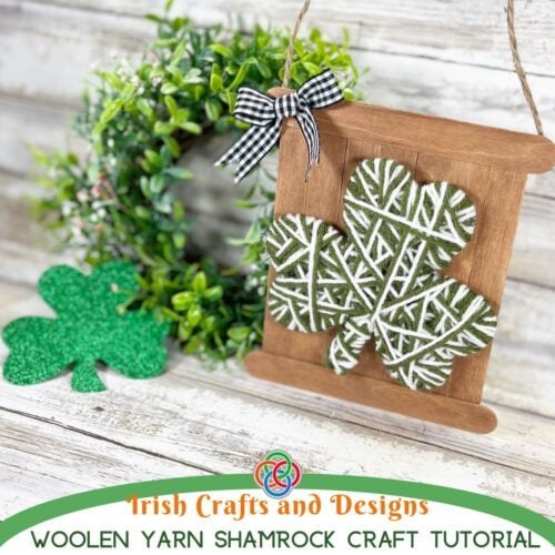 Yarn wrapped green and white shamrock mounted on a rustic craft stick sign with a gingham bow and twine hanger beside a wreath for a St. Patrick's Day craft display.