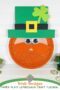 Leprechaun paper plate craft with glittery orange beard, googly eyes, green hat with yellow buckle, and shamrock decoration for a St. Patrick’s Day kids craft tutorial.