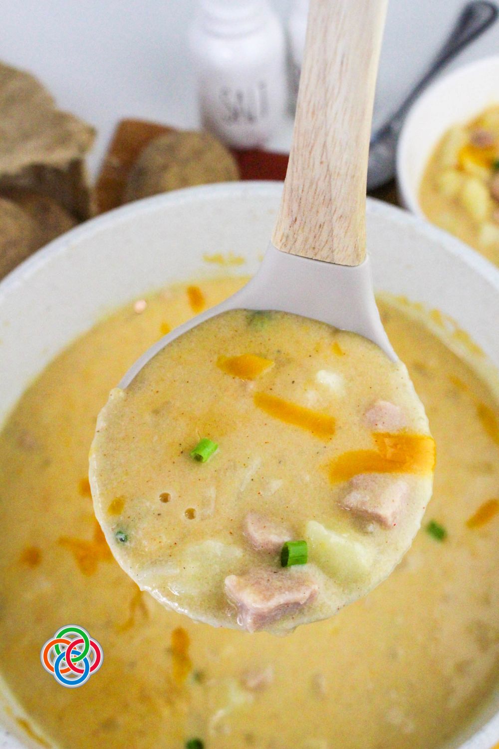 Ladle filled with creamy cheddar ham and potato soup showing chunks of potato, ham, melted cheese, and green onions in a rich, thick broth.