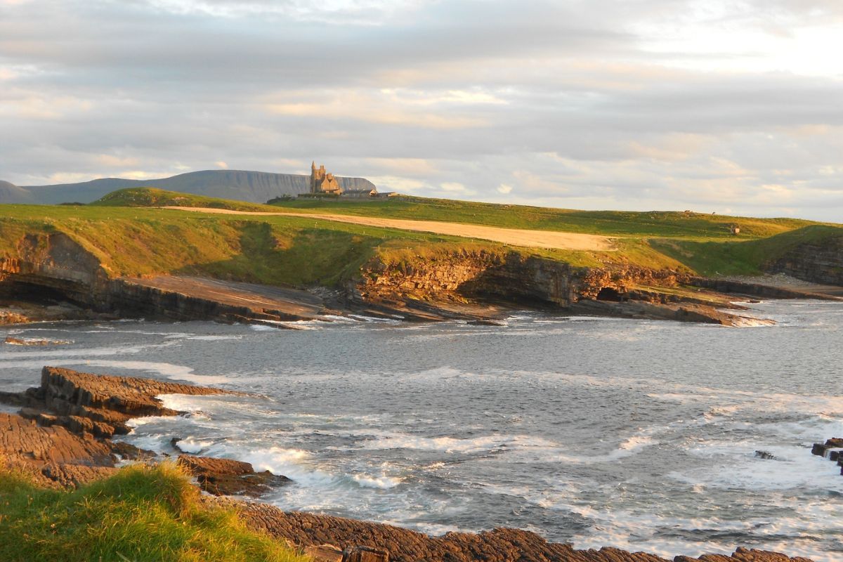 Mullaghmore in County Sligo | Irish American Mom Rocky coastline and Atlantic waves at Mullaghmore in County Sligo, Ireland, with Classiebawn Castle visible on the distant green headland under soft evening light.