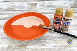 Paper plate painted with bright orange beard and flesh tone face beside acrylic paint bottles and paintbrush during a paper plate leprechaun craft tutorial.