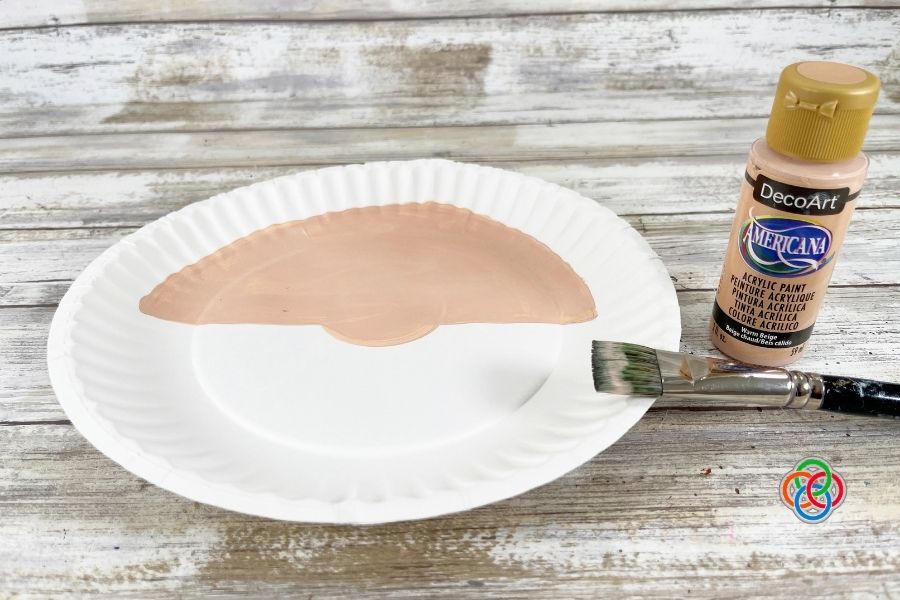Paper plate with the top half painted flesh tone for a leprechaun face beside acrylic paint and paintbrush during a paper plate leprechaun craft tutorial.
