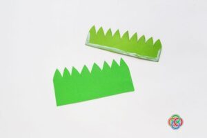 Two green paper grass cutouts with glue applied to one piece, showing how to create a layered grass pocket for a paper tulip craft.