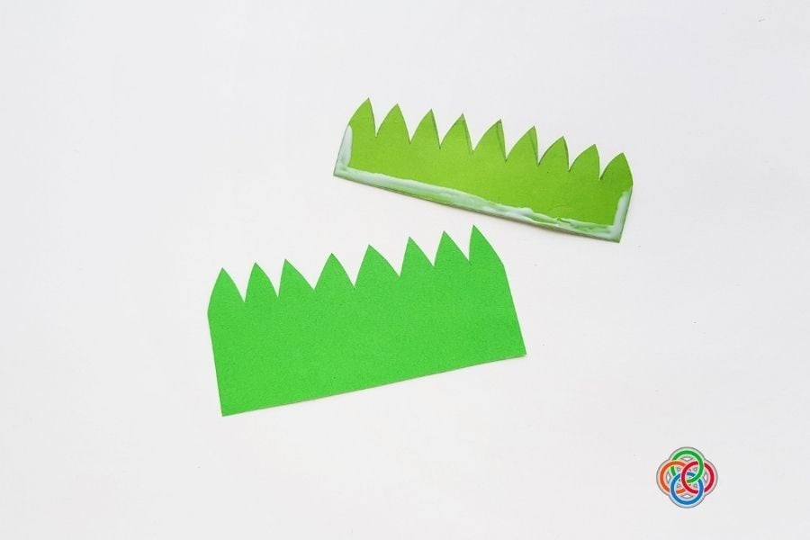 Two green paper grass cutouts with glue applied to one piece, showing how to create a layered grass pocket for a paper tulip craft.
