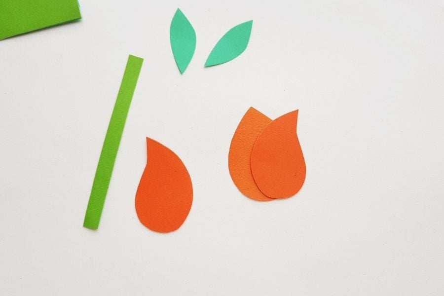 Paper tulip craft pieces including green stem and leaves with orange petals partially assembled to form a flower on a white background.