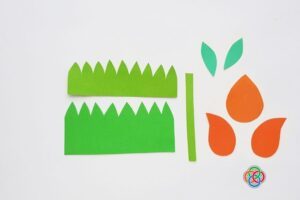 Cut-out paper pieces for a tulip craft including green grass strips, stem, leaves, and orange flower petals arranged on a white background.