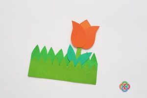 Paper tulip craft with orange flower and green leaves placed inside a layered green grass pocket on a white background.