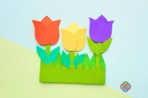 Colorful paper tulip craft with orange, yellow, and purple flowers arranged in a green grass pocket on a pastel background.