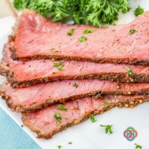 Slices of cooked corned beef brisket with a seasoned crust garnished with parsley on a white plate.