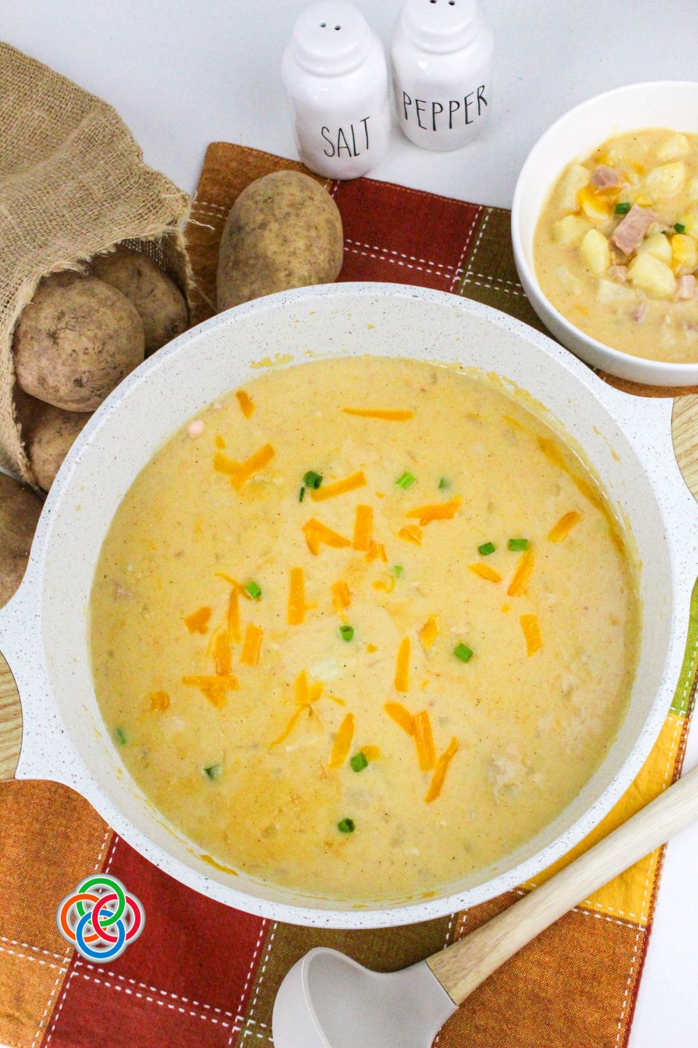 Pot of creamy cheddar ham and potato soup topped with shredded cheese and green onions, with whole potatoes and seasoning in the background.