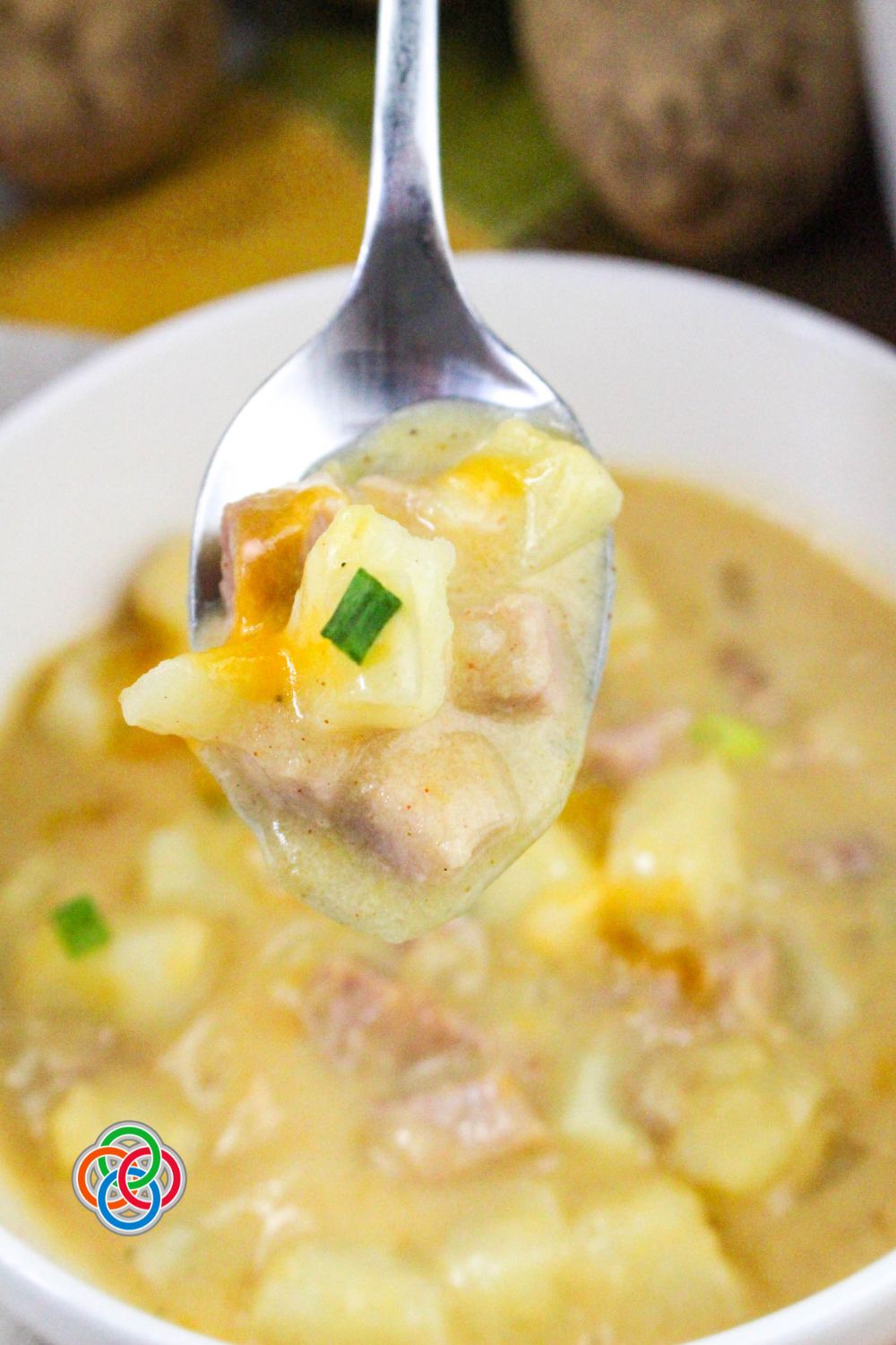 Close-up of a spoonful of creamy ham and potato soup with chunks of potato, ham, melted cheddar cheese, and green onion garnish.