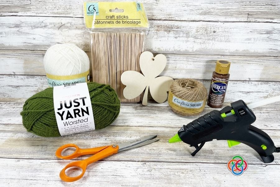 Dollar store craft supplies including yarn, craft sticks, shamrock cutout, twine, paint, scissors, and glue gun for a St. Patrick’s Day yarn wrapped shamrock sign craft.
