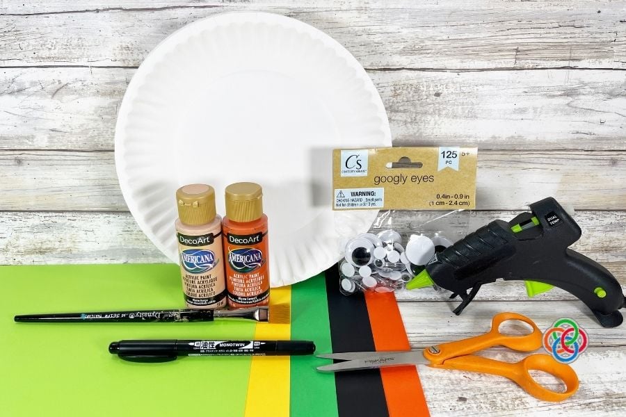 Craft supplies for a paper plate leprechaun craft including paper plate, acrylic paint, googly eyes, cardstock in green black yellow and orange, paintbrush, scissors, marker, and glue gun on a wooden background.