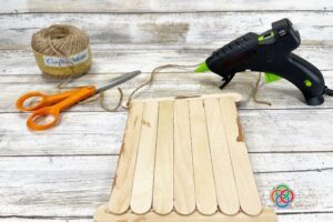 Craft stick sign with jute twine attached for hanging, alongside scissors, twine, and a glue gun during a St. Patrick's Day shamrock craft project.
