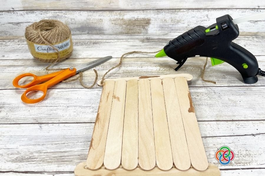 Craft stick sign with jute twine attached for hanging, alongside scissors, twine, and a glue gun during a St. Patrick’s Day shamrock craft project.