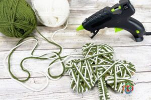 Green and white yarn wrapped around a cardboard shamrock shape with yarn skeins and a glue gun nearby for a St. Patrick's Day craft project.