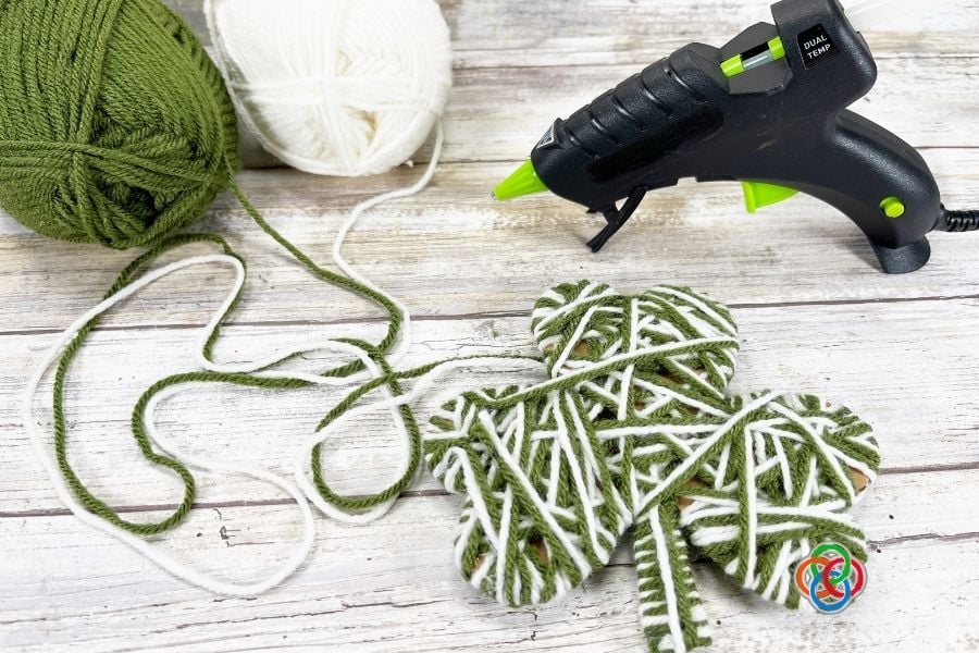 Green and white yarn wrapped around a cardboard shamrock shape with yarn skeins and a glue gun nearby for a St. Patrick’s Day craft project.