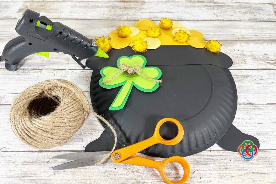 Paper plate pot of gold craft with yellow coins and pom poms, featuring a green shamrock decorated with a twine bow, with scissors, twine, and a glue gun nearby on a wooden surface.