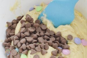 Chocolate chips and pastel M&M candies being added to soft cake mix cookie dough in a mixing bowl with a blue spatula.