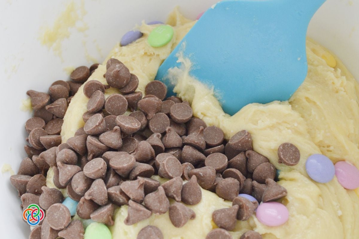Chocolate chips and pastel M&M candies being added to soft cake mix cookie dough in a mixing bowl with a blue spatula.