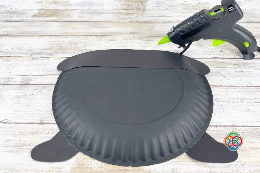Black painted paper plate shaped like a pot with attached paper legs and rim, with a glue gun nearby on a wooden surface, as part of a pot of gold craft project.