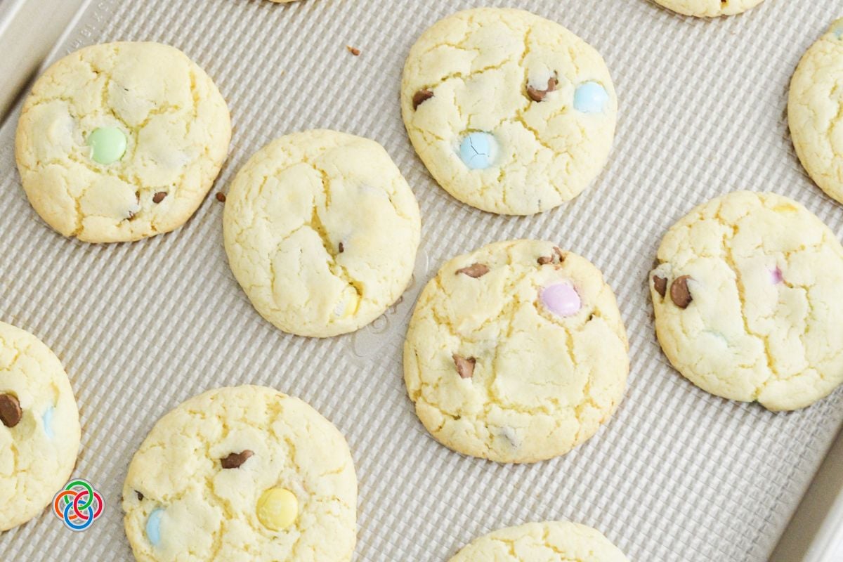 Freshly baked spring cake mix cookies with pastel M&M candies and chocolate chips cooling on a baking sheet.