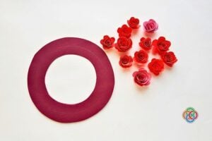 Flat lay of a large maroon circular ring beside a cluster of red paper roses on a light background, with a small multicolor logo in the bottom-right corner.