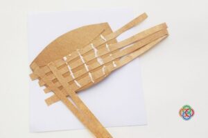 Paper Easter basket base with brown strips being woven across glued lines to create a woven pattern, with extra strips extending from the sides.