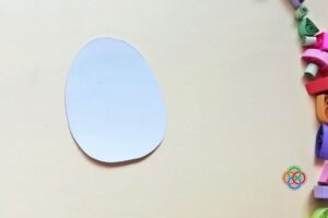White cardstock cut into an egg shape placed on a plain background with colorful quilling paper strips visible along the side.