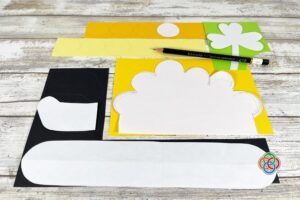 Craft supplies for a pot of gold project including cut black paper pot pieces, yellow circles for coins, a green shamrock cutout, and scissors arranged on a wooden surface.