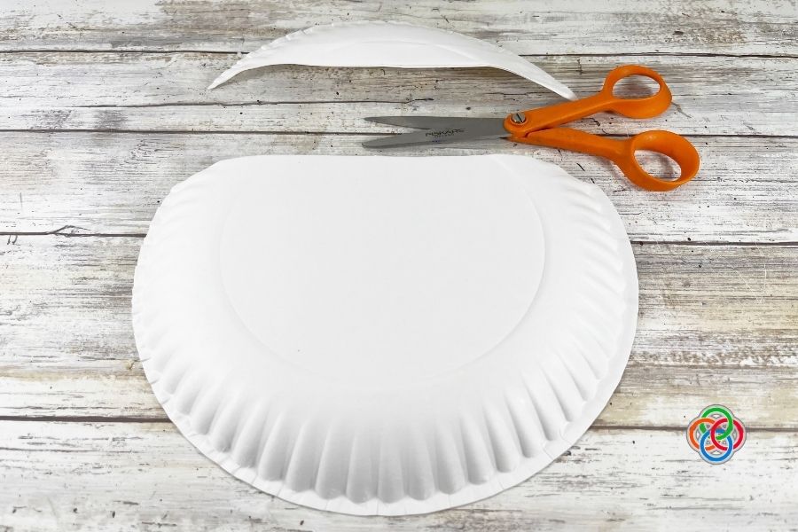 White paper plate with the top section cut off and scissors nearby on a wooden surface, showing the first step in making a pot of gold craft.