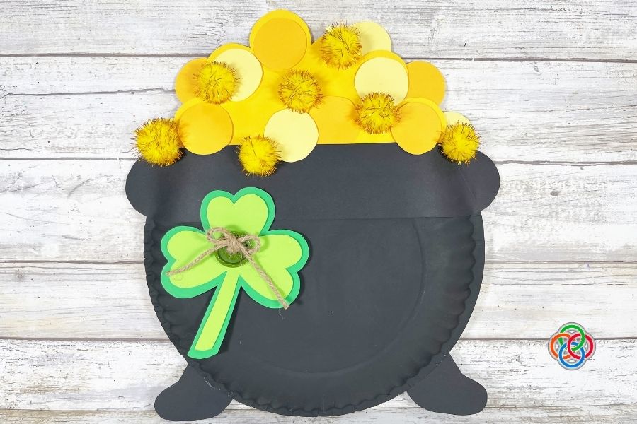 Completed paper plate pot of gold craft decorated with yellow paper coins, gold pom poms, and a green shamrock with twine, displayed on a wooden background.