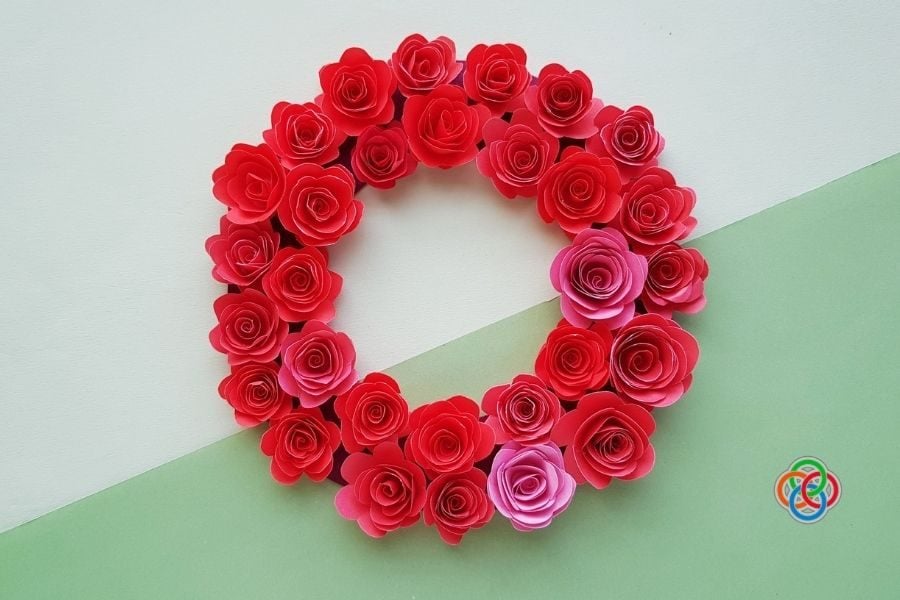 Decorative wreath of red and pink paper roses on a pale diagonal background; small multicolored logo in the bottom-right corner.