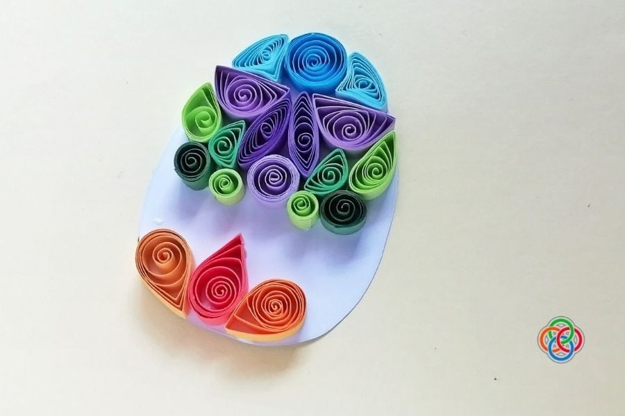 Partially completed quilled Easter egg with colorful paper shapes arranged on a white egg template, showing the design being filled in step by step.