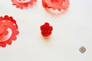 Center red paper rose surrounded by pink paper rosettes on a white surface with a small multicolor craft logo in the corner.