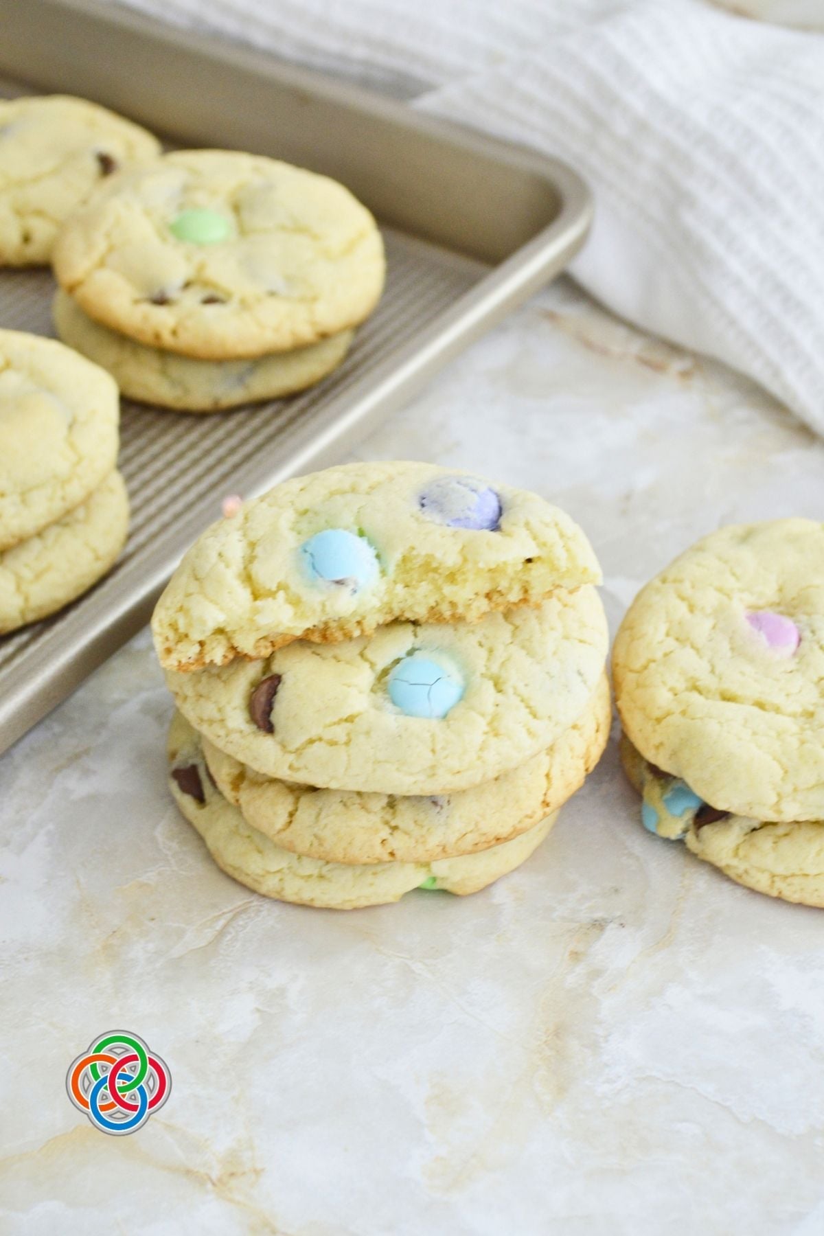 Freshly baked spring cake mix cookies stacked on a surface with pastel M&M candies and chocolate chips, with a baking tray in the background.