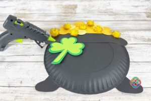 Paper plate pot of gold craft with yellow coins and gold pom poms, as a green shamrock is being glued onto the front using a glue gun on a wooden surface.