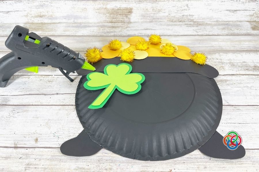 Paper plate pot of gold craft with yellow coins and gold pom poms, as a green shamrock is being glued onto the front using a glue gun on a wooden surface.