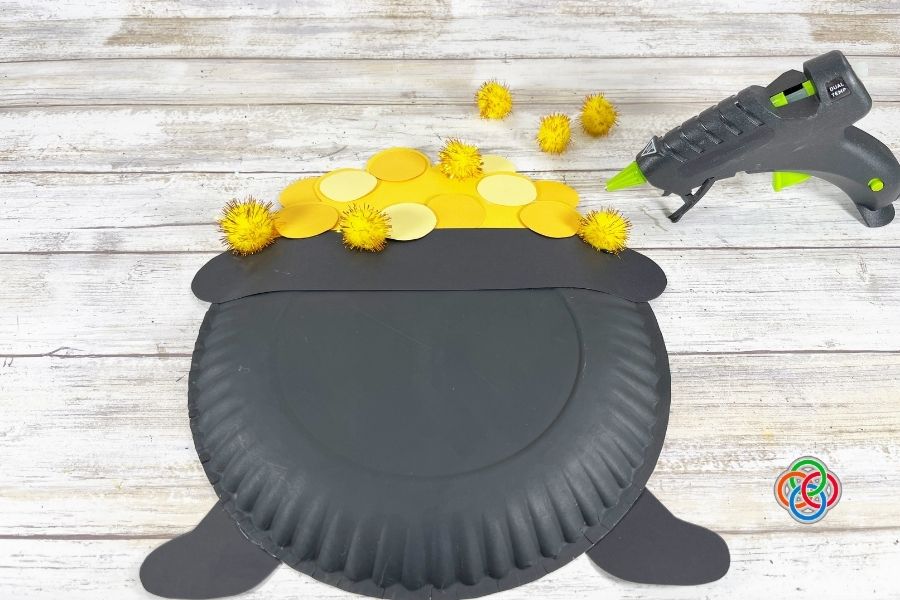 Paper plate pot of gold craft with yellow paper coins and gold pom poms being added on top, with a glue gun nearby on a wooden surface.