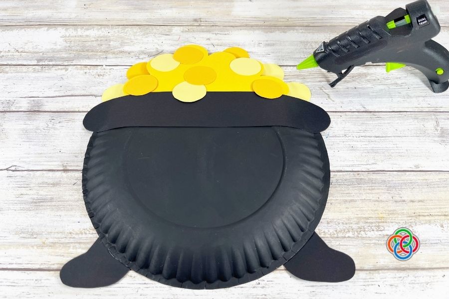 Paper plate pot of gold craft with yellow paper circles glued on top to resemble coins, with a glue gun nearby on a wooden surface.