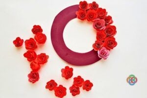 Maroon circular wreath partially covered with red paper roses, with loose petals scattered on a white surface.