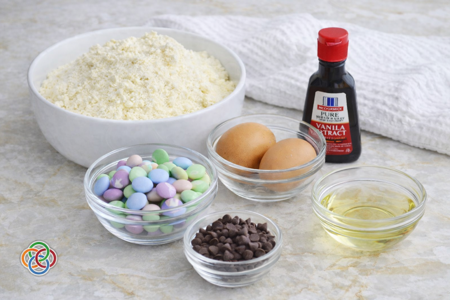 Ingredients for cake mix cookies including yellow cake mix, eggs, vegetable oil, vanilla extract, chocolate chips, and pastel M&M candies arranged in bowls on a marble surface.