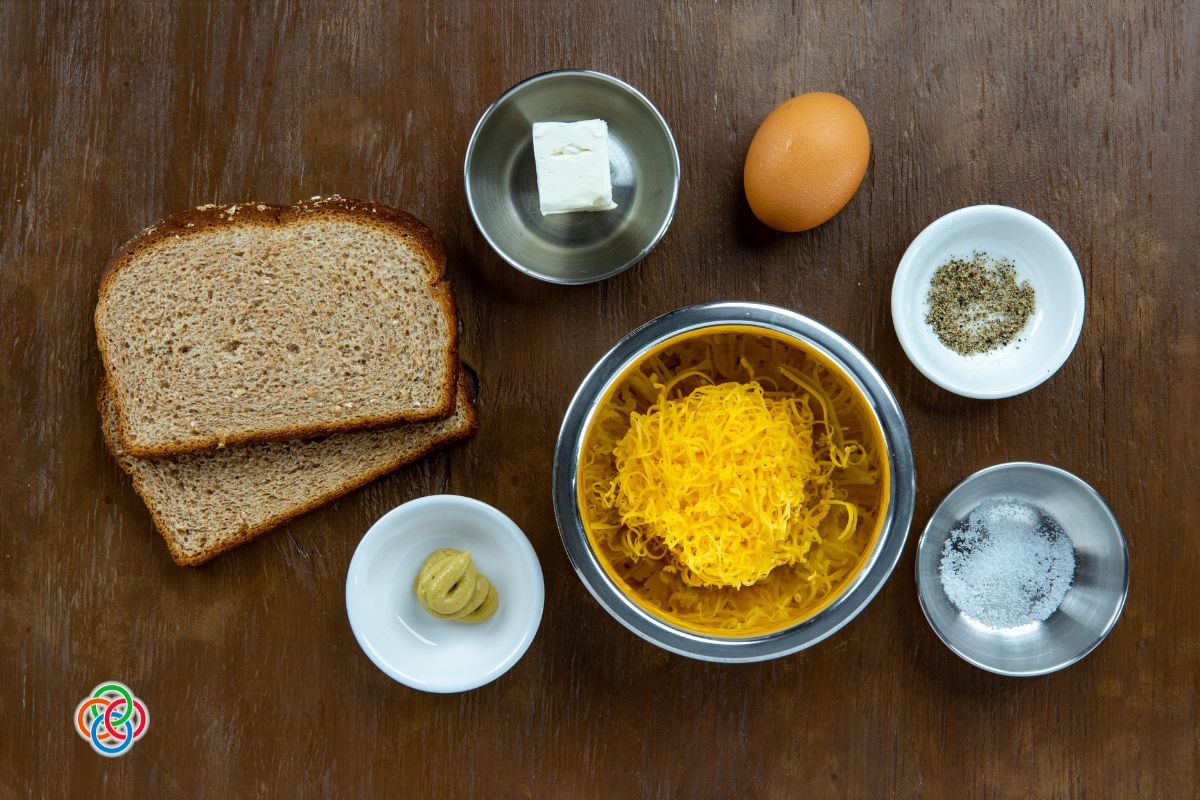 Top-down view of cooking prep: two slices of bread, a pat of butter, an egg, salt, pepper, and a bowl of macaroni with shredded cheddar on a wooden surface.