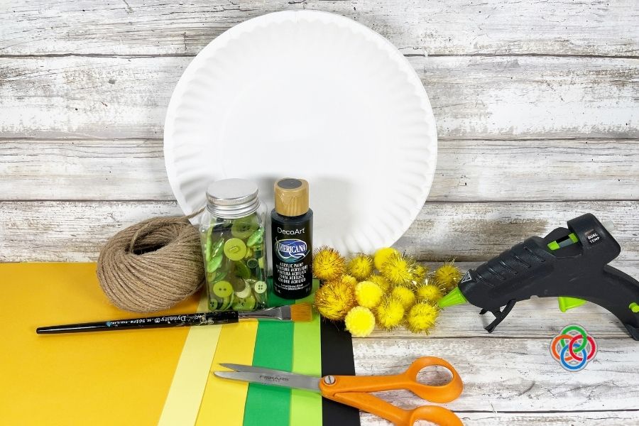 Craft materials for a pot of gold project including a paper plate, black paint, glue gun, scissors, colored paper, pom-poms, twine, and buttons arranged on a wooden surface.