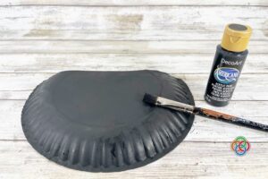Paper plate being painted black with a brush and acrylic paint bottle on a wooden surface, as part of a pot of gold craft project.