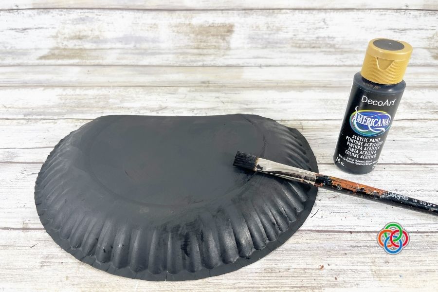 Paper plate being painted black with a brush and acrylic paint bottle on a wooden surface, as part of a pot of gold craft project.