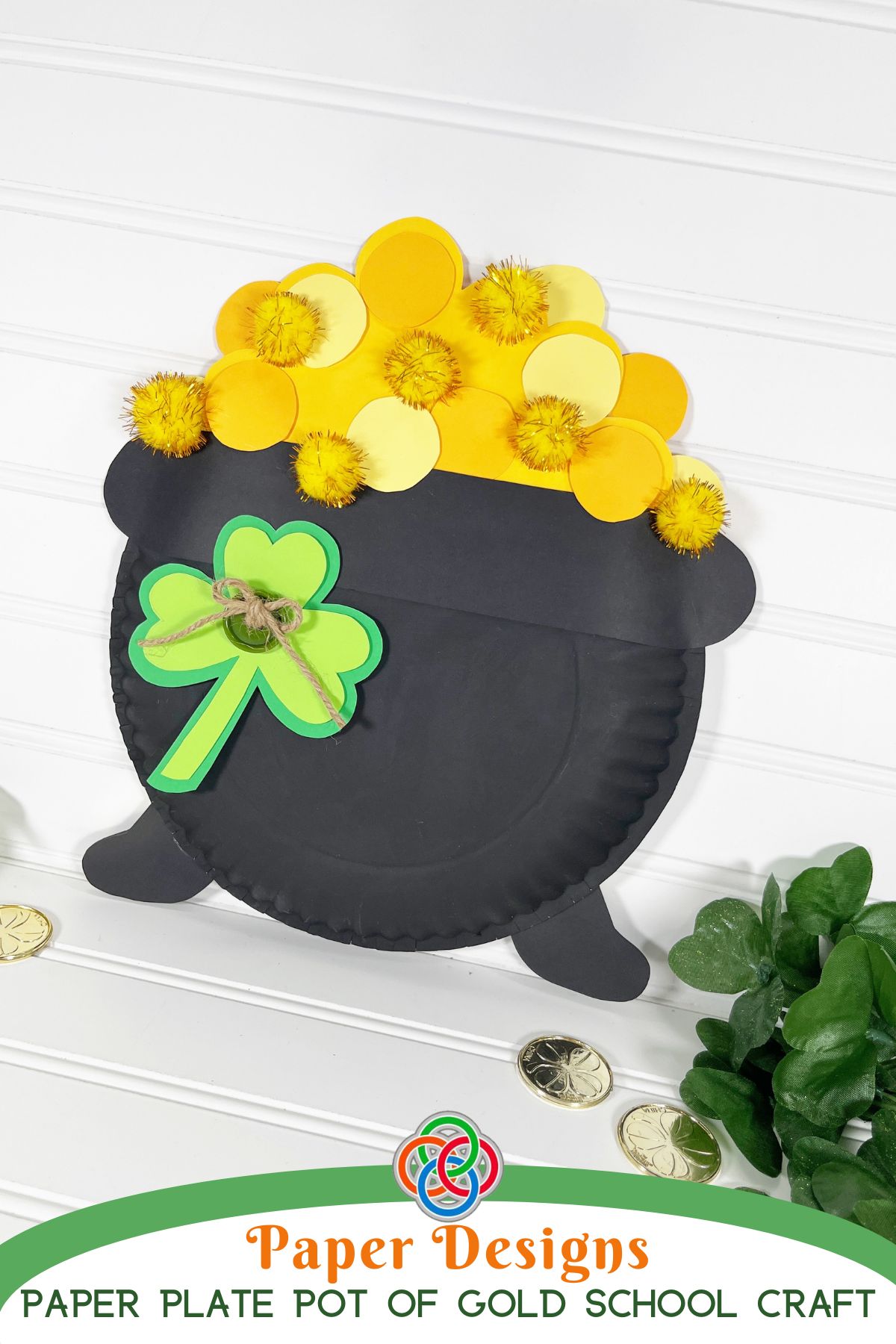 Paper plate craft shaped like a pot of gold filled with yellow paper circles and pom-poms, decorated with a green shamrock, displayed on a white background with gold coins and greenery, with text reading “Paper Plate Pot of Gold School Craft.”