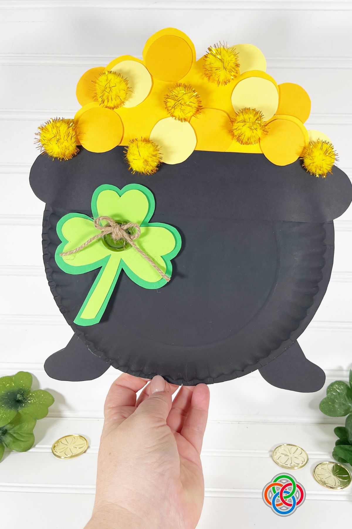 Hand holding a paper plate craft shaped like a pot of gold filled with yellow paper circles and pom-poms, decorated with a green shamrock, on a white background with scattered gold coins.
