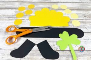 Craft supplies for a pot of gold project including cut black paper pot pieces, yellow circles for coins, a green shamrock cutout, and scissors arranged on a wooden surface.