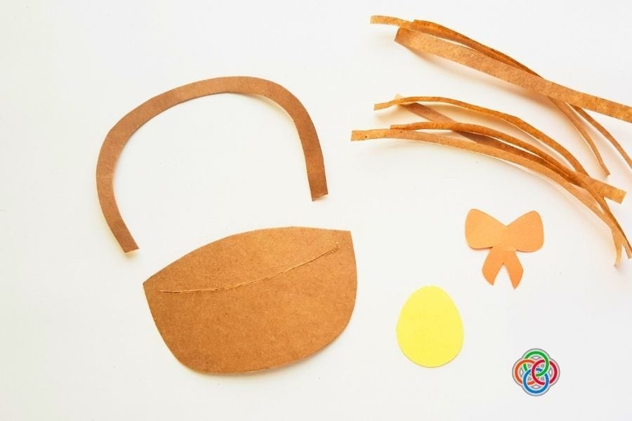 Cut-out paper pieces for an Easter basket craft, including basket base, handle, strips, bow, and egg shape, arranged on a plain background.